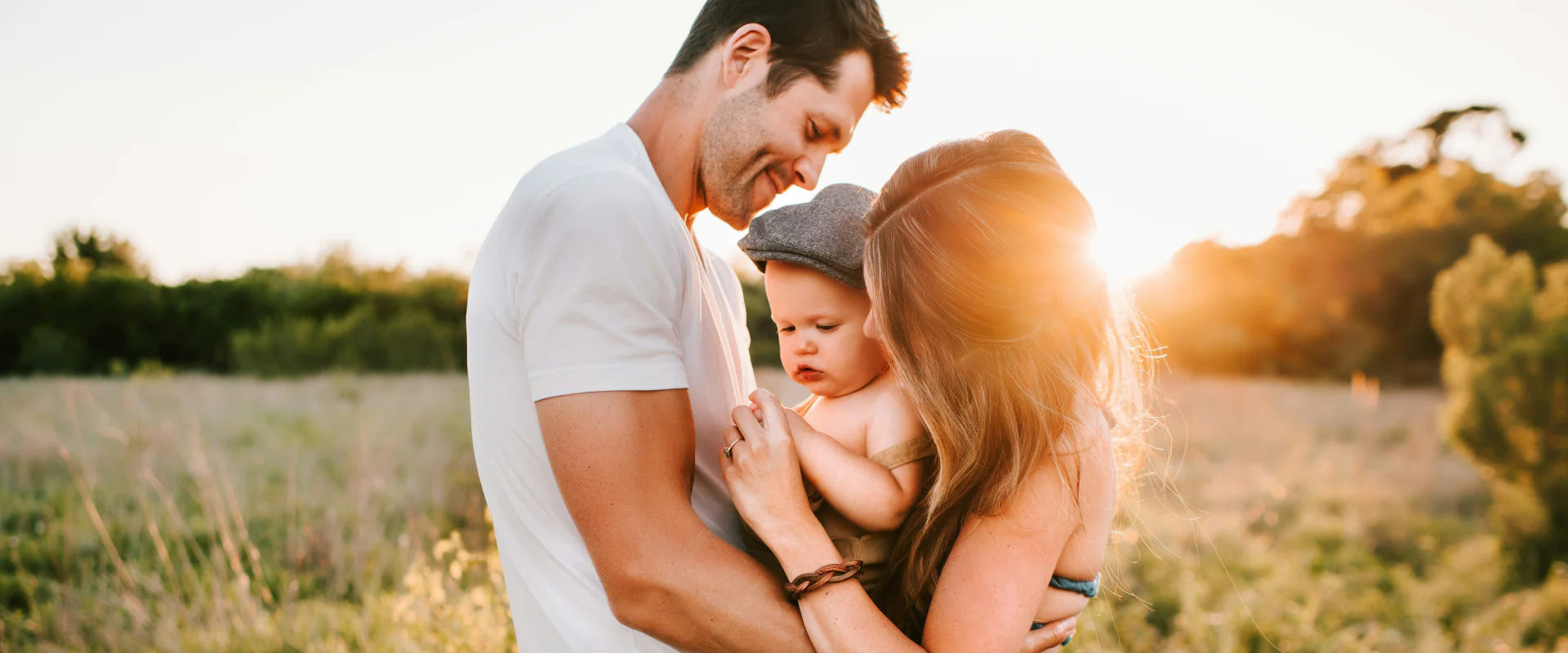 Eltern halten ihr Baby im Arm, auf einer Wiese im Abendlicht