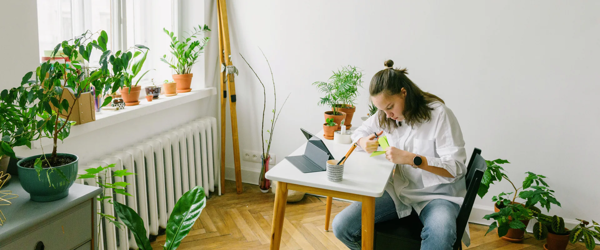 Junge Frau macht sich am Schreibtisch in einem hellen Zimmer voller Pflanzen Notizen