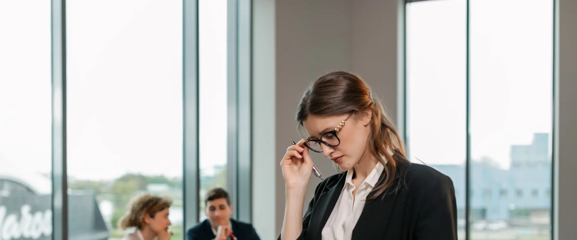 Managerin mit Brille liest Unterlagen, im Hintergrund eine Besprechung am Konferenztisch