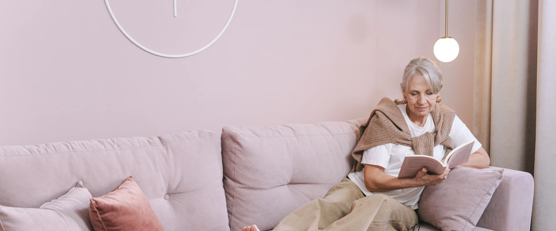 Ältere Frau liest entspannt auf einem rosa Sofa in ihrer hellen Wohnung