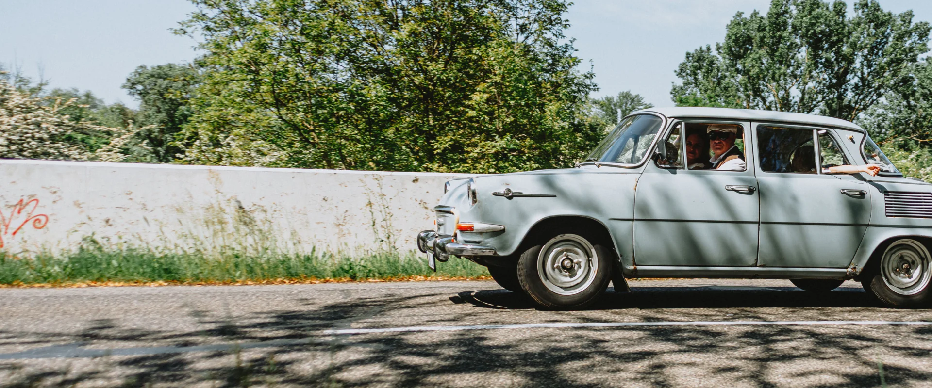 Heller Oldtimer mit Fahrer auf einer sonnigen Straße vor Bäumen