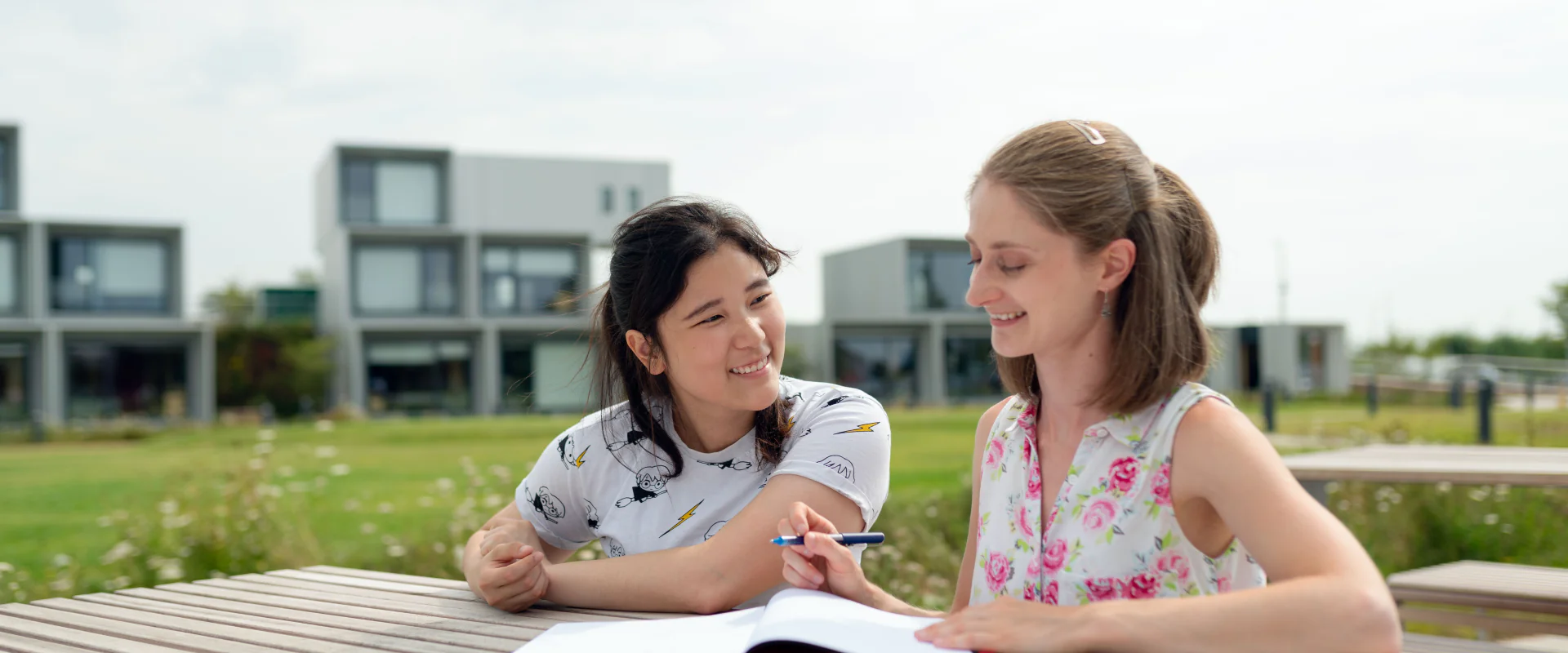 Zwei Studentinnen lernen lachend an einem Tisch auf dem Campus
